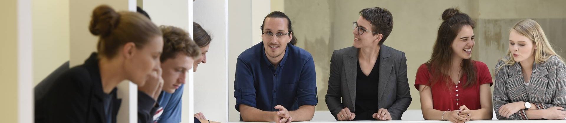 Eine Gruppe von Menschen blickt auf eine Brüstung gelehnt nach unten, einige unterhalten sich.