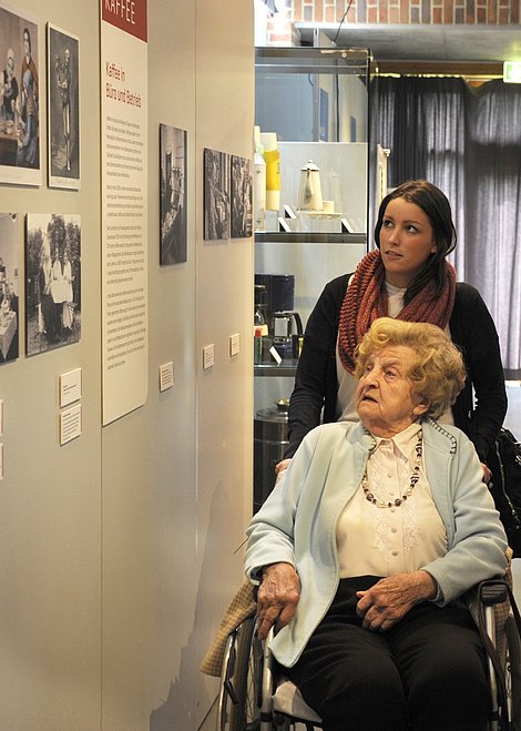 Eine junge Frau schiebt eine ältere Frau im Rollstuhl durch eine Ausstellung.