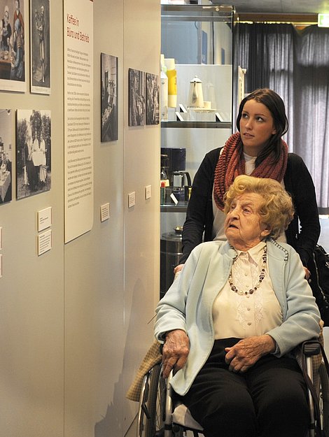 Eine junge Frau schiebt eine ältere Frau im Rollstuhl durch eine Ausstellung.