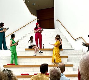 Auf einer Treppen stehen mehrere Menschen, die jeweils in einer Farbe gekleidet sind: rot, grün, gelb, blau, schwarz und weiß. Sie sprechen miteinander. Unten an der Treppe hochblickend sieht Publikum ihnen zu.
