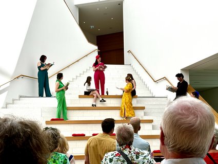 Auf einer Treppen stehen mehrere Menschen, die jeweils in einer Farbe gekleidet sind: rot, grün, gelb, blau, schwarz und weiß. Sie sprechen miteinander. Unten an der Treppe hochblickend sieht Publikum ihnen zu.