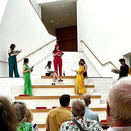 Auf einer Treppen stehen mehrere Menschen, die jeweils in einer Farbe gekleidet sind: rot, grün, gelb, blau, schwarz und weiß. Sie sprechen miteinander. Unten an der Treppe hochblickend sieht Publikum ihnen zu.